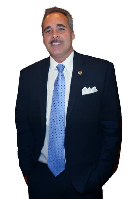 John Fleming - A man in a dark suit with a light blue tie and white pocket square stands facing the camera with hands in his pockets, smiling slightly—an image evocative of the professional poise of Home John M. Fleming. - Charlotte County Commissioner