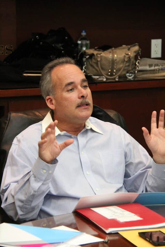 John Fleming - About John M. Fleming: A man in a light blue shirt sits at a desk with colorful folders, gesturing with both hands while speaking. - Charlotte County Commissioner