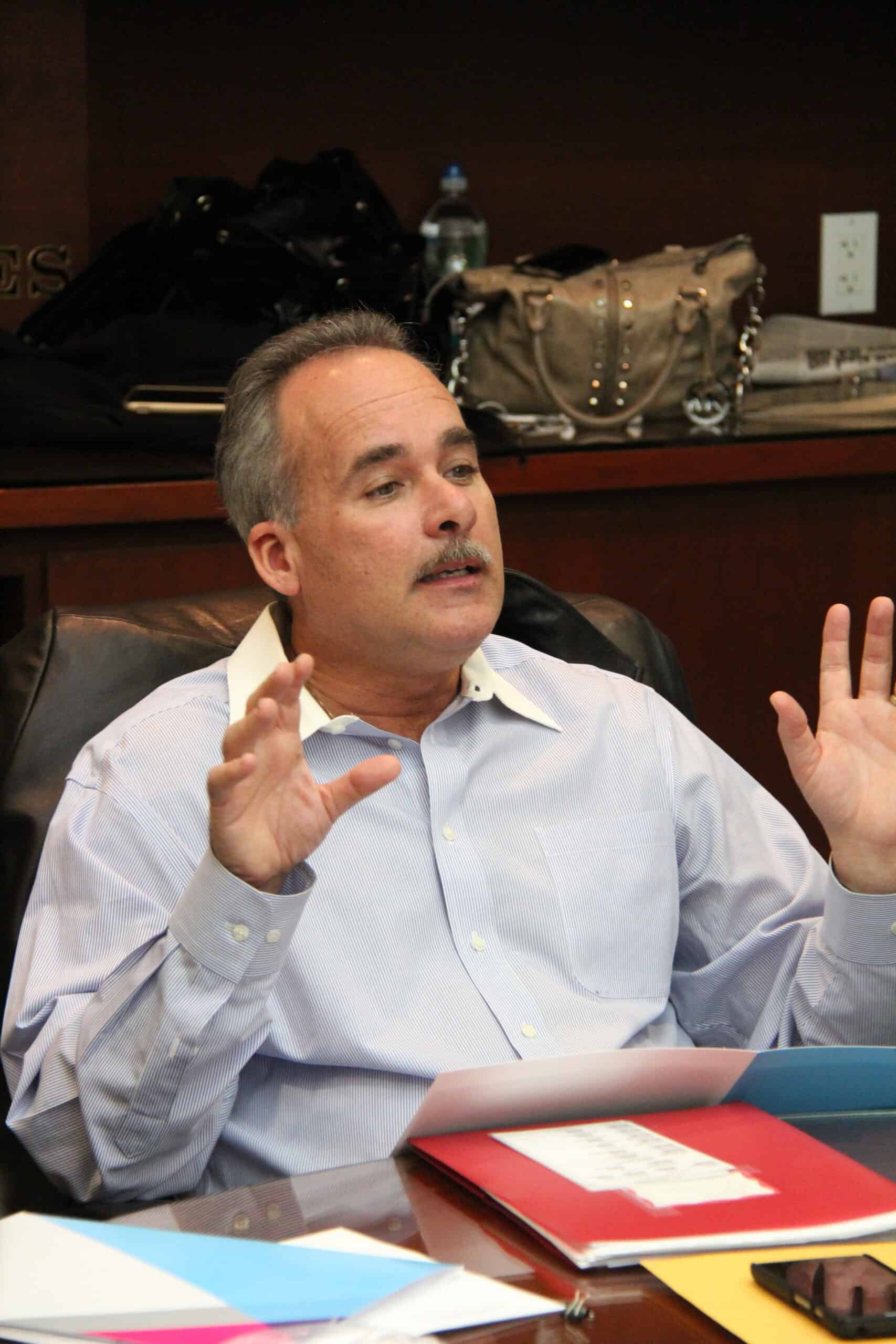 John Fleming - About John M. Fleming: A man in a light blue shirt sits at a desk with colorful folders, gesturing with both hands while speaking. - Charlotte County Commissioner