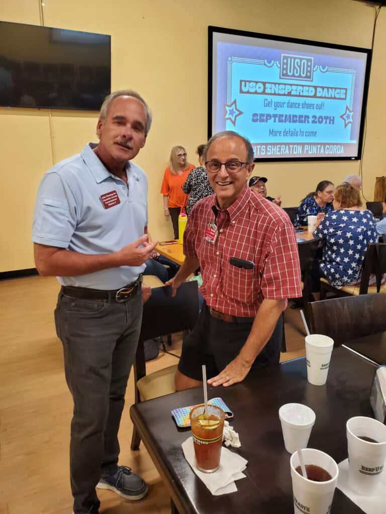 John Fleming - Two men pose and smile at a table during an indoor event, with people seated in the background and an event announcement about John M. Fleming projected on the wall. - Charlotte County Commissioner