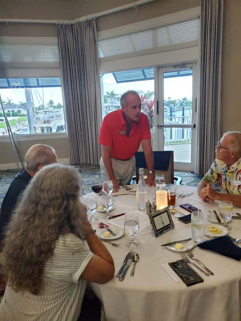 John Fleming - About John M. Fleming: A man in a red shirt stands and speaks to three seated people at a round dining table set with food and drinks in a restaurant with large windows. - Charlotte County Commissioner
