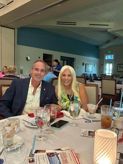 John Fleming - A man and woman sit together at a dining table in a restaurant, smiling for the camera. The table has drinks, glasses, brochures, and a candle. Other people are visible in the background, including About John M. Fleming brochures on the table. - Charlotte County Commissioner